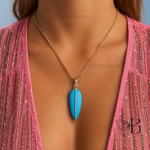 May include: A gold-toned necklace featuring a pendant with a vibrant turquoise feather design. The feather pendant has detailed carved lines and a gold accent. The necklace is displayed against a pink background.