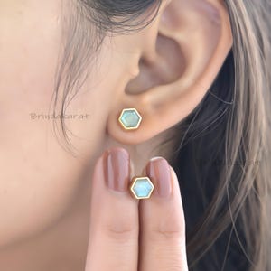 May include: Hexagonal stud earrings with a gold border and a pale blue-green centre stone. One earring is in the ear, and the other is held by a finger. The earrings are from Brindakarat.