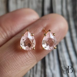 May include: A pair of gold stud earrings with pear-shaped, faceted, pale pink gemstones. Each earring has a gold post and is accented with small gold beads. The earrings are shown against a blurred background.