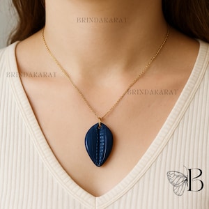 May include: A gold-colored chain necklace with a dark blue pendant. The pendant is an oval shape with a textured, ribbed design. The necklace is worn against a cream-colored top.