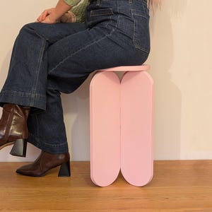 Könnte beinhalten: Ein hellrosa, moderner Hocker mit einem einzigartigen, geschwungenen Design. Der Hocker hat eine kreisförmige Oberseite und zwei gebogene, vertikale Stützen. Die Person, die auf dem Hocker sitzt, trägt eine dunkelblaue Jeans und braune Stiefel.