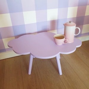 May include: A lavender cloud-shaped side table with three legs. A pink teacup and a white and pink gradient coffee pot sit on top. The table is made of wood and is painted a solid lavender color.