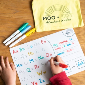 May include: A white dry-erase board with a colorful alphabet printed on it. A child is writing on the board with a red dry-erase marker. The board also has a section for writing a name and age. The text on the board reads "my name is Lake. I am 6 years old."  The board is labeled "MOO + DOT Adventures in color".