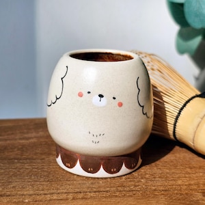 Könnte beinhalten: Beige Keramik-Matcha-Schale mit einem Cartoon-Bärengesicht-Design. Die Schale hat einen braunen Innenraum und steht auf einem wellenförmigen braunen Sockel. Ein Bambusbesen ist im Hintergrund zu sehen.