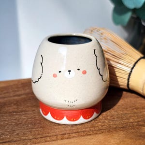 Könnte beinhalten: Beige Keramikbecher mit einem Bärengesicht-Design. Der Becher hat einen roten, gewellten Boden und eine dunkelblaue Innenseite. Schwarze Linien umreißen die Ohren und das Gesicht des Bären. Ein Bambusbesen ist im Hintergrund zu sehen.
