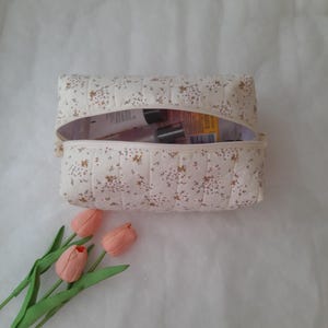 K&ouml;nnte beinhalten: Eine rechteckige, gesteppte Kosmetiktasche aus cremefarbenem Stoff mit einem kleinen braun-gelben Blumenmuster. Die Tasche hat eine Rei&szlig;verschluss&ouml;ffnung an der Oberseite, die mehrere darin aufbewahrte Flaschen zeigt, und wird von drei pfirsichfarbenen Tulpen begleitet.