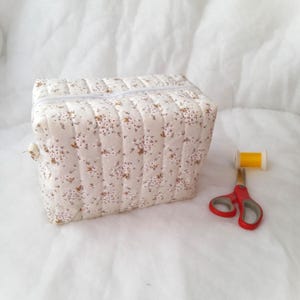 May include: A quilted, rectangular beige fabric case with a floral pattern and a white zipper. A spool of yellow thread and red scissors are next to the case. The case is likely for sewing supplies.