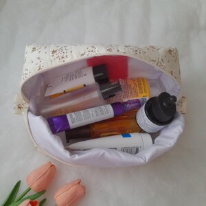 K&ouml;nnte beinhalten: Ein gesteppter Stoffbeutel mit cremefarbener Au&szlig;enseite und kleinem Blumenmuster, gef&uuml;llt mit verschiedenen Hautpflegeflaschen und Tuben. Das Innere ist mit wei&szlig;em Stoff ausgekleidet und der Beutel ist offen, um die Kosmetikbeh&auml;lter zu zeigen.