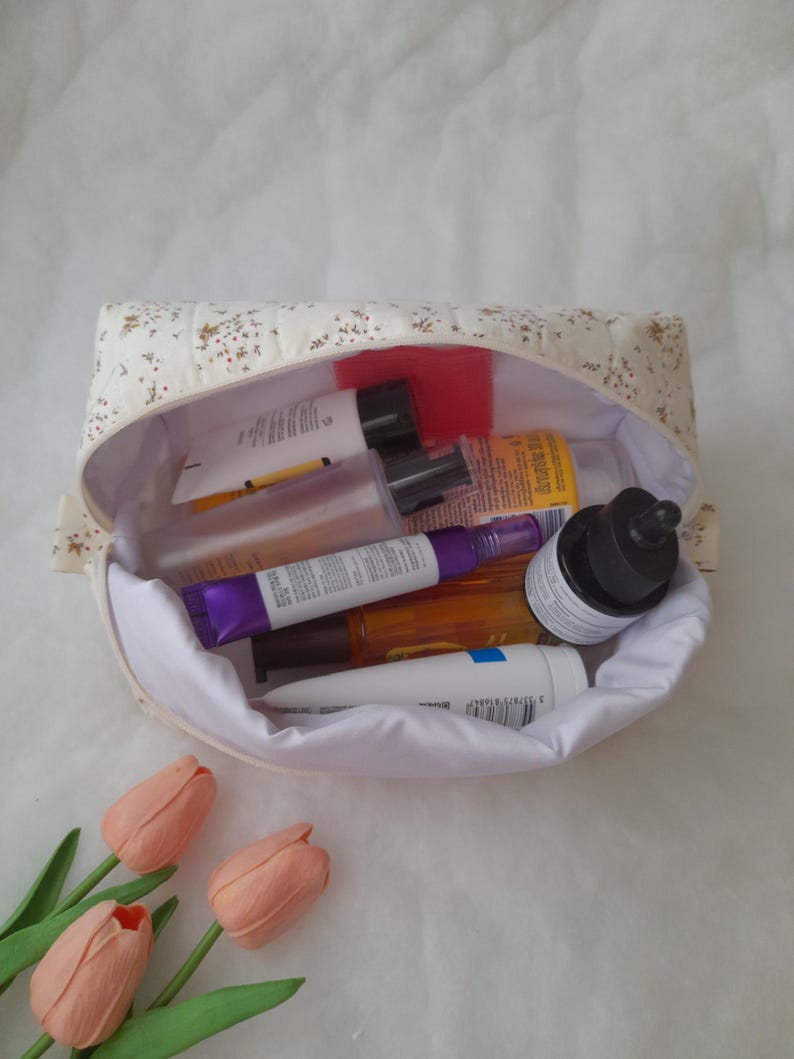 K&ouml;nnte beinhalten: Ein gesteppter Stoffbeutel mit cremefarbener Au&szlig;enseite und kleinem Blumenmuster, gef&uuml;llt mit verschiedenen Hautpflegeflaschen und Tuben. Das Innere ist mit wei&szlig;em Stoff ausgekleidet und der Beutel ist offen, um die Kosmetikbeh&auml;lter zu zeigen.