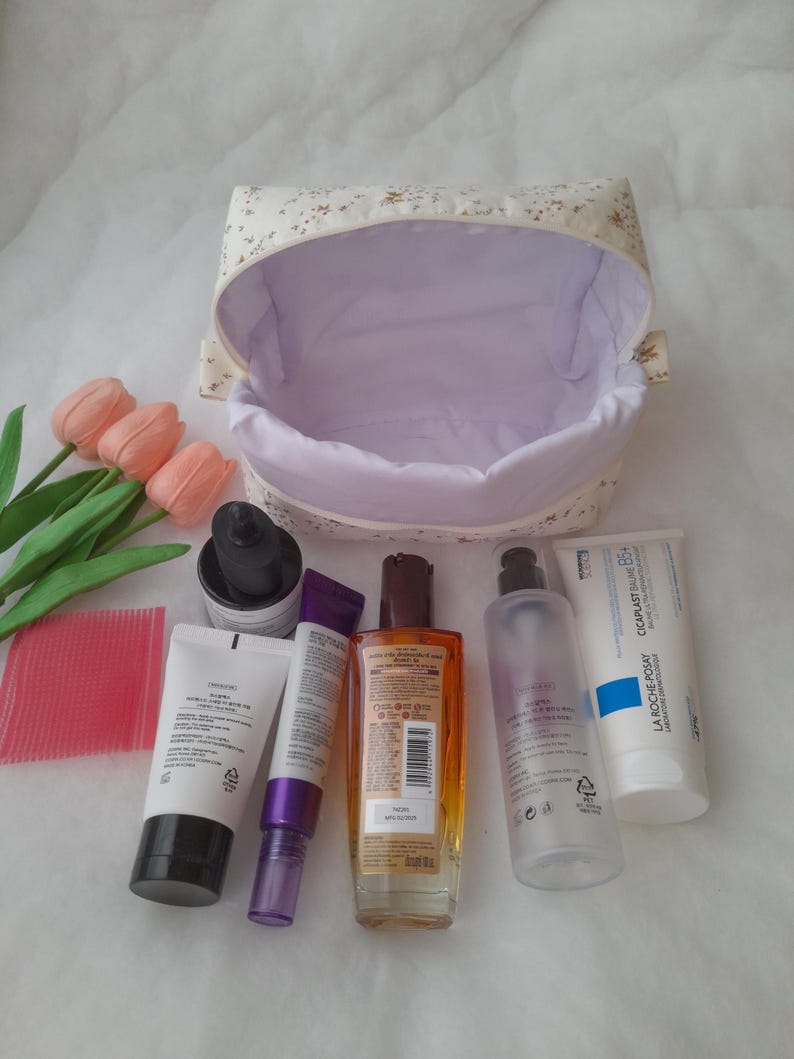 K&ouml;nnte beinhalten: Eine Kosmetiktasche aus Stoff mit Blumenmuster und hellviolettem Innenfutter, begleitet von drei k&uuml;nstlichen rosa Tulpen, einem rosa Kamm sowie sechs verschiedenen Hautpflegeflaschen und -tuben, darunter eine La Roche-Posay Cicaplast Baume B5+ Tube.