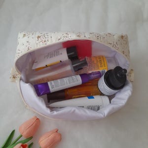 K&ouml;nnte beinhalten: Ein gesteppter Stoffbeutel mit cremefarbener Au&szlig;enseite und kleinem Blumenmuster, gef&uuml;llt mit verschiedenen Hautpflegeflaschen und Tuben. Das Innere ist mit wei&szlig;em Stoff ausgekleidet und der Beutel ist offen, um die Kosmetikbeh&auml;lter zu zeigen.