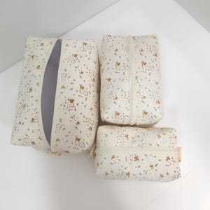 K&ouml;nnte beinhalten: Ein Set aus drei rechteckigen Rei&szlig;verschlusstaschen in verschiedenen Gr&ouml;&szlig;en aus cremefarbenem Stoff mit einem zarten Blumenmuster aus winzigen braunen und wei&szlig;en Bl&uuml;ten. Jede Tasche hat einen passenden cremefarbenen Rei&szlig;verschluss und eine gesteppte Textur.