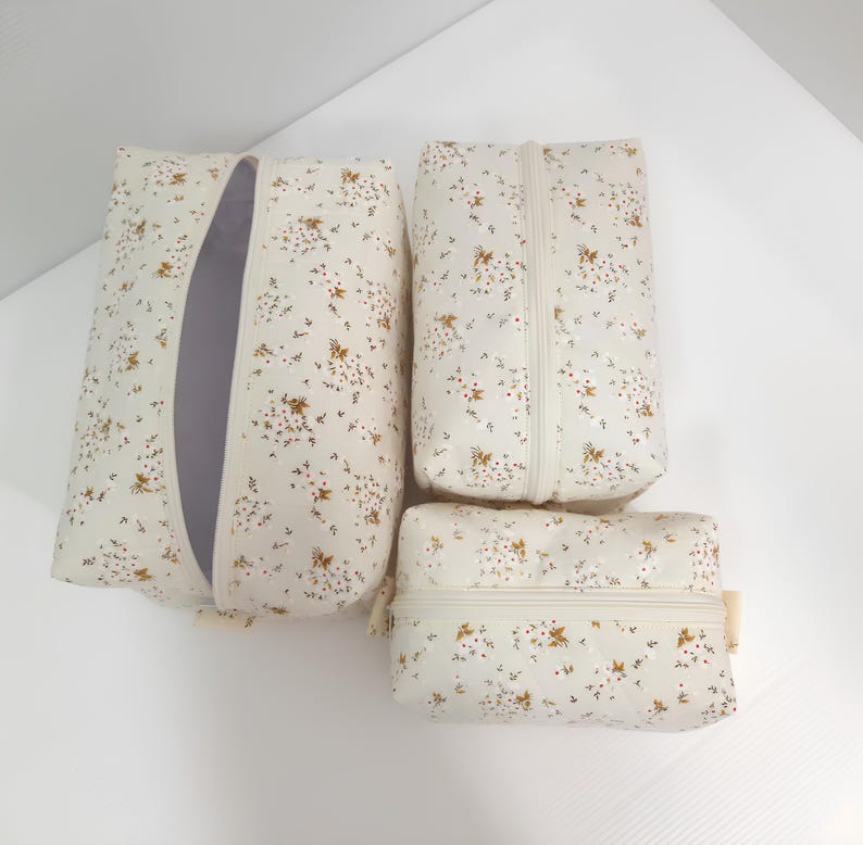 K&ouml;nnte beinhalten: Ein Set aus drei rechteckigen Rei&szlig;verschlusstaschen in verschiedenen Gr&ouml;&szlig;en aus cremefarbenem Stoff mit einem zarten Blumenmuster aus winzigen braunen und wei&szlig;en Bl&uuml;ten. Jede Tasche hat einen passenden cremefarbenen Rei&szlig;verschluss und eine gesteppte Textur.