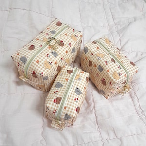 Può includere: Tre trousse per cosmetici a quadretti beige e bianchi con un motivo ripetuto di orsetti, ciliegie e fiori. Ogni trousse ha una cerniera dorata e un ciondolo decorativo.