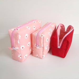 May include: Three zippered pouches, one red and two pink with a white daisy print. The pouches are lined up in order of size, with the largest pouch on the left and the smallest pouch on the right.