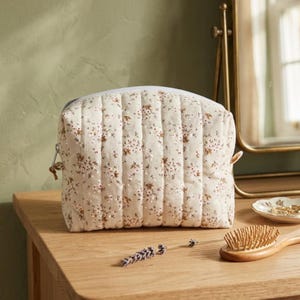 May include: A quilted pouch with a cream-colored base featuring a small-scale floral pattern in shades of brown and white. The pouch has a rectangular shape with vertical stitching lines and a silver-toned zipper closure along the top edge.