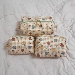 K&ouml;nnte beinhalten: Drei gesteppte Taschen in verschiedenen Gr&ouml;&szlig;en mit beigefarbenem und wei&szlig;em Vichy-Muster, verziert mit kleinen braunen und grauen B&auml;ren, roten Kirschen und orangefarbenen Blumen. Jede Tasche hat einen Rei&szlig;verschluss mit goldenen blumenf&ouml;rmigen Anh&auml;ngern.