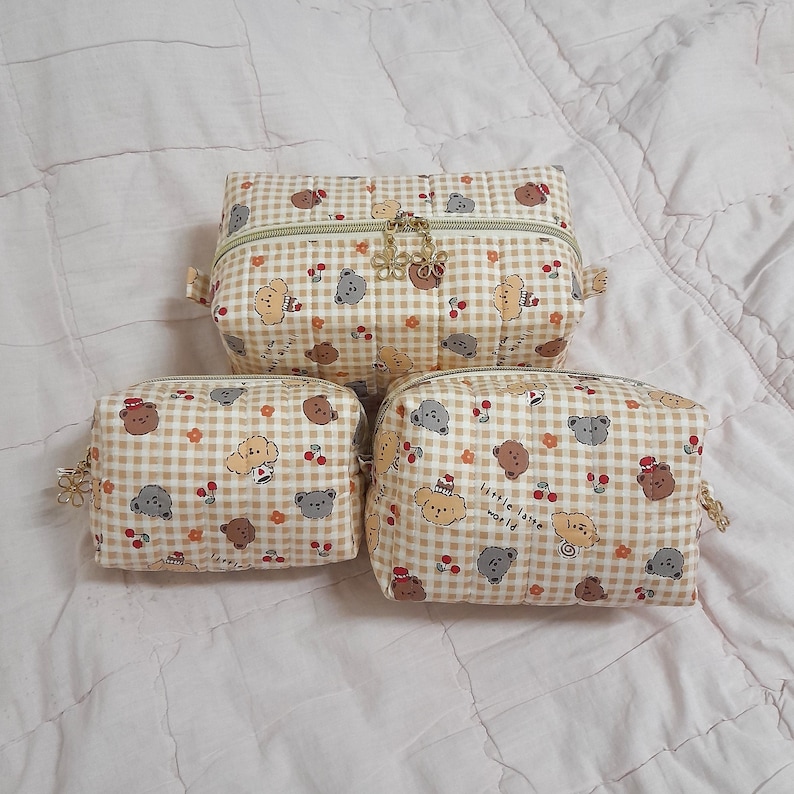K&ouml;nnte beinhalten: Drei gesteppte Taschen in verschiedenen Gr&ouml;&szlig;en mit beigefarbenem und wei&szlig;em Vichy-Muster, verziert mit kleinen braunen und grauen B&auml;ren, roten Kirschen und orangefarbenen Blumen. Jede Tasche hat einen Rei&szlig;verschluss mit goldenen blumenf&ouml;rmigen Anh&auml;ngern.
