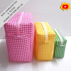 Può includere: Tre astucci rettangolari con cerniera di dimensioni decrescenti, caratterizzati da un motivo a quadretti vichy rosa, giallo e verde. Ogni astuccio ha una cerniera in tinta sul bordo superiore. Il testo sull'immagine recita 100% HANDCRAFTED & TESTED SEWING PATTERN (NO AI).
