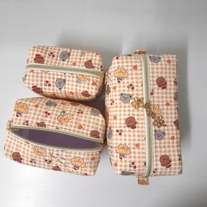 K&ouml;nnte beinhalten: Drei gesteppte Stofftaschen in verschiedenen Gr&ouml;&szlig;en mit beigefarbenem und wei&szlig;em Vichy-Muster, verziert mit kleinen Illustrationen von braunen und grauen B&auml;ren sowie roten Kirschen. Jede Tasche hat einen hellgr&uuml;nen Rei&szlig;verschluss und einen Blumenanh&auml;nger.