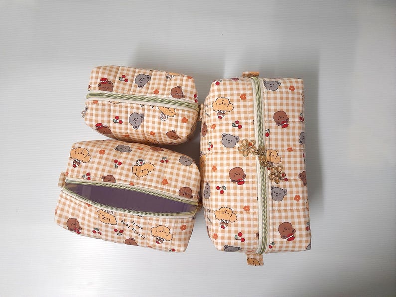 K&ouml;nnte beinhalten: Drei gesteppte Stofftaschen in verschiedenen Gr&ouml;&szlig;en mit beigefarbenem und wei&szlig;em Vichy-Muster, verziert mit kleinen Illustrationen von braunen und grauen B&auml;ren sowie roten Kirschen. Jede Tasche hat einen hellgr&uuml;nen Rei&szlig;verschluss und einen Blumenanh&auml;nger.