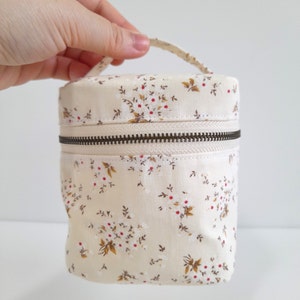 May include: A cylindrical cream-colored fabric bag with a floral pattern of white flowers and red berries. The bag has a dark brown zipper and a small handle. The bag is approximately 5 inches tall and 3 inches in diameter.