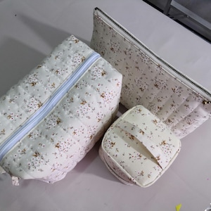 May include: A set of three quilted cosmetic bags in varying sizes. The bags are off-white with a delicate floral pattern in brown and red. Each bag has a zipper closure, with a light blue zipper on the largest bag. The smallest bag is square with a handle.