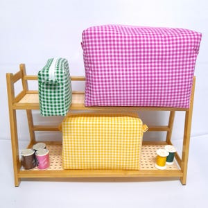 Könnte beinhalten: Ein Holzregal zeigt drei Taschen mit Vichy-Karo-Muster in Grün, Gelb und Pink. Auf den Regalen befinden sich auch Garnrollen in verschiedenen Farben und kleine Behälter. Die Taschen sind rechteckig und haben einen Reißverschluss.