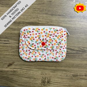 May include: A rectangular fabric pouch featuring a white background with a repeating pattern of small orange and red flowers with green stems. The pouch has a front flap secured by a single red snap button and a white zipper closure along the top edge.