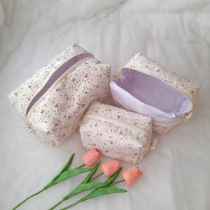 K&ouml;nnte beinhalten: Drei gesteppte Kosmetiktaschen in verschiedenen Gr&ouml;&szlig;en aus cremefarbenem Stoff mit einem zarten braunen Blumenmuster. Jede Tasche hat einen Rei&szlig;verschluss und ein einfarbig wei&szlig;es Innenfutter. Drei k&uuml;nstliche pfirsichfarbene Tulpen liegen darunter.