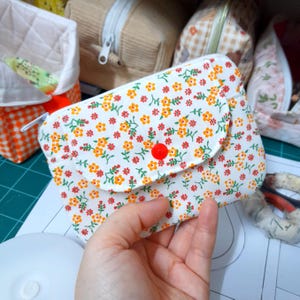 Könnte beinhalten: Eine kleine, weiße Tasche mit floralem Muster und rotem Knopfverschluss. Die Tasche hat ein Muster aus orangefarbenen und gelben Blumen mit grünen Blättern. Die Tasche wird in einer Hand gehalten.