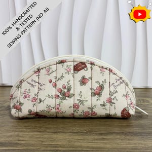 May include: A semi-circular quilted pouch featuring a cream-colored fabric with a repeating pattern of small pink flowers, green leaves, and brown baskets. The pouch has a white zipper closure along the top edge and vertical quilted stitching lines.