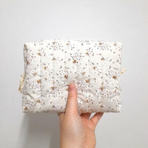 May include: A quilted, rectangular cosmetic bag with a white background and a floral pattern in brown and white. The bag is held by a hand and has a zipper closure. The bag has a soft, padded appearance.