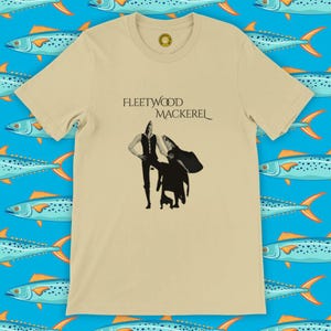 May include: A beige t-shirt with the words "FLEETWOOD MACKEREL" printed above a black and white illustration of two figures. The tee has a crew neck and short sleeves. A small circular logo is on the upper chest.