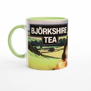 Könnte beinhalten: Weißer Keramikbecher mit grünem Henkel und Rand. Der Becher zeigt den Text "BJORKSHIRE TEA" in einem schwarzen Rechteck, mit einer ländlichen Szene mit grünen Hügeln und einer Frau.
