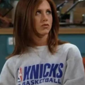 May include: A light gray long-sleeve crewneck sweatshirt featuring the blue text KNICKS and the word BASKETBALL printed below it, accompanied by a red, white, and blue NBA logo graphic on the left side of the chest area.