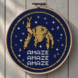 Peut inclure: Broderie au point de croix dans un cercle en bois. Le motif présente une figure abstraite pixélisée dans des tons de beige, de vert et de rose sur un fond bleu foncé. Le mot « AMAZE » est répété trois fois en blanc. Le cercle est fixé avec un fermoir en métal.