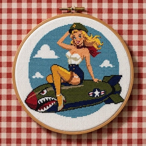 May include: Cross-stitch embroidery in a wooden hoop. The design features a blonde woman in a military-style hat and outfit, seated on a bomb. The background is blue with white clouds. The bomb is green with a red nose.