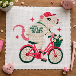 May include: A cross-stitch embroidery on white fabric featuring a white rat wearing a red cap and riding a pink bicycle. The rat has a long pink tail and a green strap. The text reads: DONT ANALIZE LIFE MORE THAN YOU LIVE IT! A green basket with white flowers is attached to the handlebars.