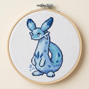 May include: A cross-stitch embroidery hoop featuring a blue creature with large ears and a long tail. The creature is stitched in shades of blue and white on a white fabric background, set in a wooden hoop.