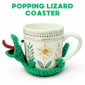 May include: A white ceramic mug with a floral design is embraced by a green crocheted lizard coaster. The lizard has a red tongue and a white eye. The text "POPPING LIZARD COASTER" is above the mug.