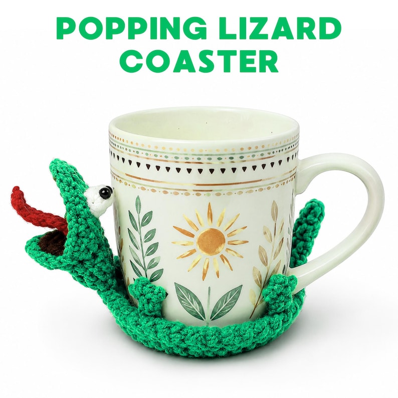 May include: A white ceramic mug with a floral design is embraced by a green crocheted lizard coaster. The lizard has a red tongue and a white eye. The text "POPPING LIZARD COASTER" is above the mug.