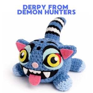 May include: A blue crocheted plush toy with large, expressive eyes, a pink tongue, and the text "DERPY FROM DEMON HUNTERS" in blue. The toy has a striped tail and pink accents on its ears.