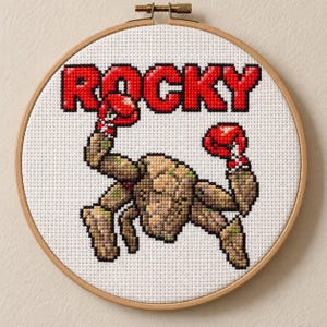 Könnte beinhalten: Eine Kreuzstichstickerei in einem runden Holzrahmen, die eine pixelige, beige, felsartige Figur mit roten Boxhandschuhen zeigt. Über der Figur ist das Wort ROCKY in fetten, roten, pixeligen Großbuchstaben auf weißem Stoff gestickt.