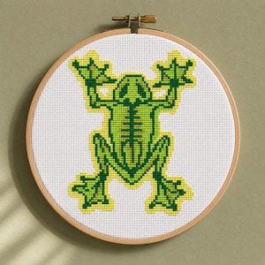 May include: A cross-stitch embroidery hoop featuring a green frog design with yellow accents on a white background. The frog is centered, with its arms and legs outstretched. The wooden hoop is round and has a brass adjustment screw.