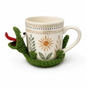 May include: A cream-colored ceramic mug with a floral design, featuring a crocheted green alligator wrapped around it. The alligator has a red tongue and white eyes. The mug has a handle and is decorated with a sun and leaf pattern.