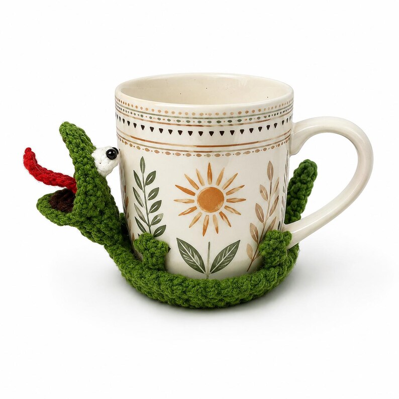 May include: A cream-colored ceramic mug with a floral design, featuring a crocheted green alligator wrapped around it. The alligator has a red tongue and white eyes. The mug has a handle and is decorated with a sun and leaf pattern.