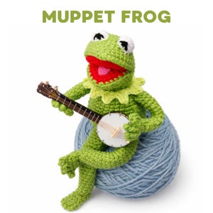 May include: A crocheted Kermit the Frog doll, holding a banjo, sits atop a blue yarn ball. The frog is green with a red mouth, white eyes, and a yellow collar. The text "MUPPET FROG" is displayed above the frog.