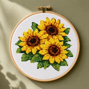 Può includere: Un telaio da ricamo circolare in legno contenente un motivo a punto croce di tre girasoli con petali giallo brillante, centri marrone scuro e foglie verde intenso, ricamati su uno sfondo di tessuto bianco con trama a griglia.