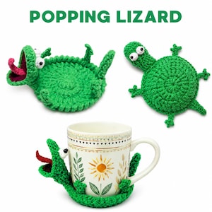 Könnte beinhalten: Drei grüne gehäkelte Eidechsen-Tassenuntersetzer und -manschetten. Eine Eidechse ist um eine weiße Tasse mit floralem Design gewickelt. Die Eidechsen haben weiße Augen und rosa Zungen. Der Text "POPPING LIZARD" befindet sich oben im Bild.